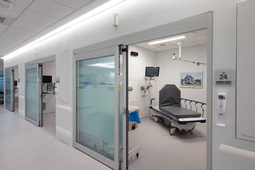 Modern hospital corridor featuring glass sliding doors open to an emergency department room with a patient bed