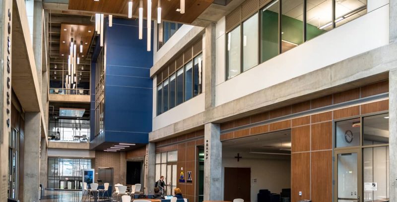 Modern multi-story building atrium with hanging pendant lights, wooden ceiling panels, and ground-floor seating